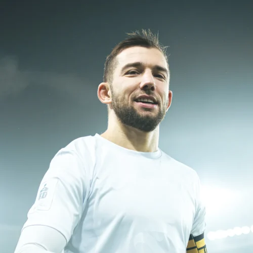 Na zdjęciu znajduje się mężczyzna w białej koszulce, uśmiechający się i patrzący w stronę obiektywu, w otoczeniu jasnego tła stadionu.