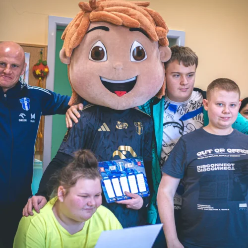 Grupa osób z uśmiechami, w tym osoba w dużej maskotce, stoi w pomieszczeniu, trzymając plany lekcji oraz kartki papieru.