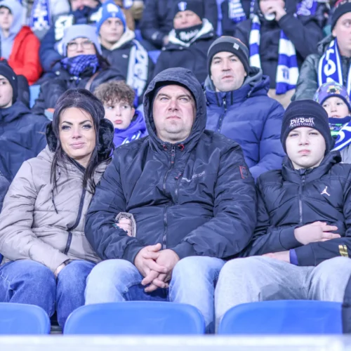 Na zdjęciu widać rodzinę siedzącą na trybunach, kibicującą w zimowej scenerii na meczu sportowym.