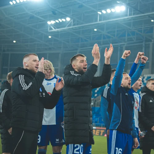 Piłkarze drużyny, ubrani w dresy i kurtki, entuzjastycznie świętują zwycięstwo, klaskając i podnosząc ręce na tle stadionu.