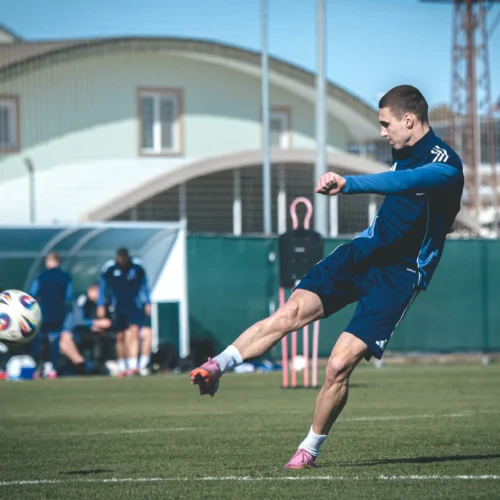 Na zdjęciu widać piłkarza wykonującego strzał na boisku treningowym, w tle znajdują się inni zawodnicy oraz infrastruktura sportowa.