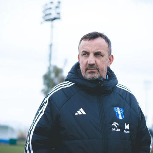 Na zdjęciu widoczny jest mężczyzna w czarnej kurtce sportowej, patrzący w dal na boisku piłkarskim, w tle znajduje się oświetlenie stadionu.