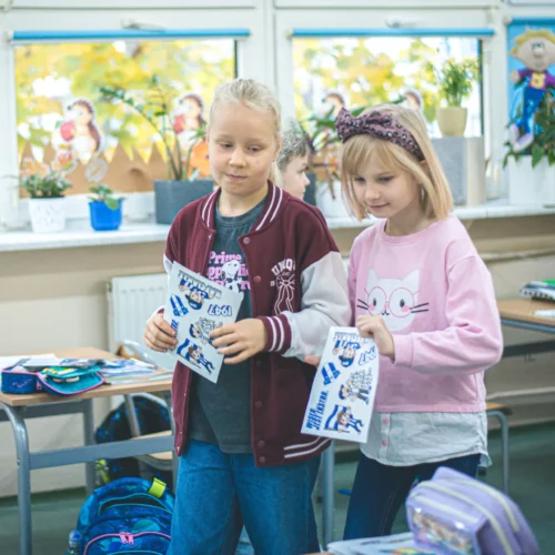 Dwie dziewczynki w szkolnej klasie trzymają kartki z rysunkami, uśmiechając się do siebie.