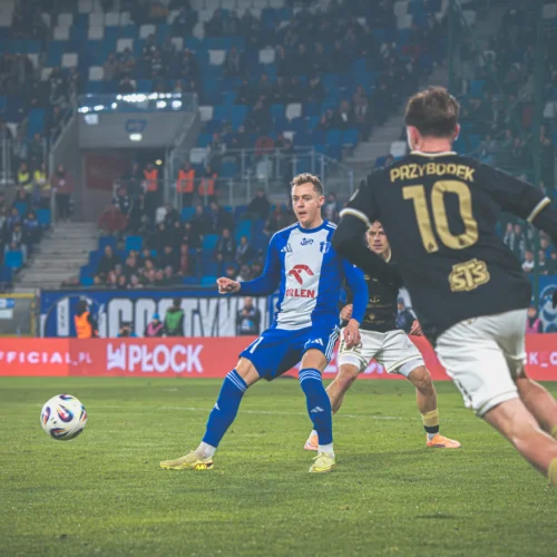 Na zdjęciu widoczna jest sytuacja podczas meczu piłkarskiego, gdzie zawodnik w niebieskim stroju kopie piłkę, a obok niego dwóch rywali w czarnych koszulkach.