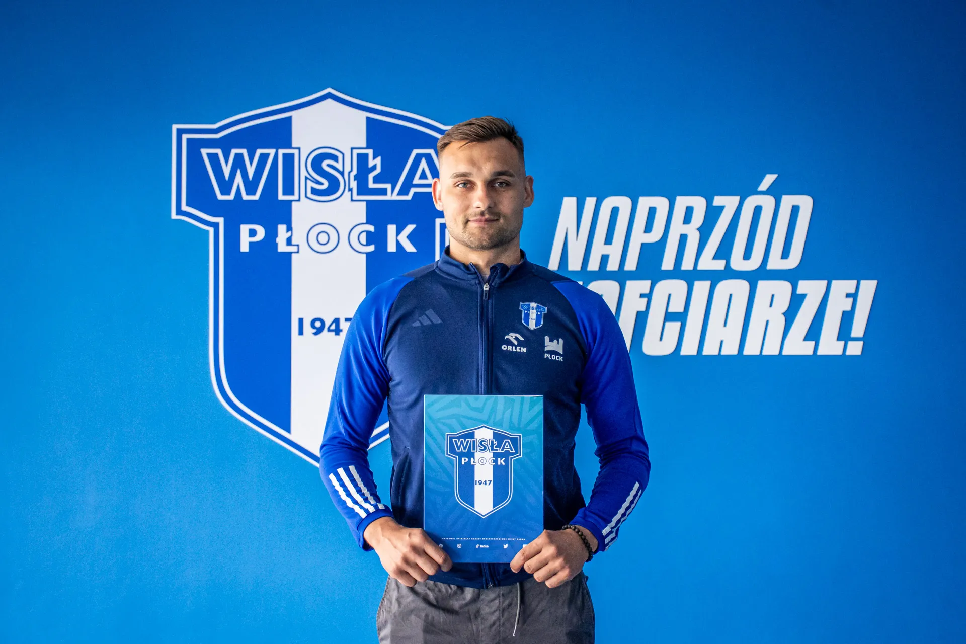 Na zdjęciu widoczny jest mężczyzna w niebieskiej koszulce sportowej, trzymający dokument w rękach, stojący przed tłem z logo klubu Wisła Płock.