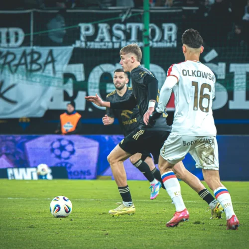 Na zdjęciu widać zawodników grających w piłkę nożną podczas meczu na boisku, z wyraźnymi transparentami w tle.