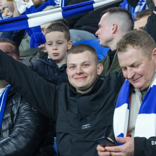 Na zdjęciu widać grupę kibiców na stadionie, którzy radośnie trzymają w górze biało-niebieskie szaliki, a wśród nich uśmiechnięty mężczyzna.