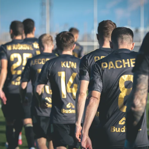 Na zdjęciu widoczna jest grupa piłkarzy w czarnych strojach z złotymi numerami, idących w kierunku boiska.