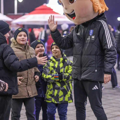 Na zdjęciu widać grupkę dzieci, które uśmiechają się i pozdrawiają osobę przebrana w kostium maskotki z dużą, uśmiechniętą twarzą.