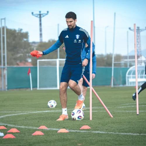 Na zdjęciu widać zawodników trenujących na boisku piłkarskim, z geometrycznymi przeszkodami i piłkami w otoczeniu zielonej murawy.