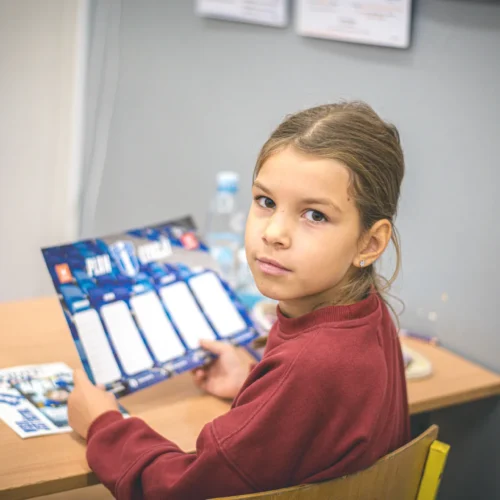 Na zdjęciu widoczna jest dziewczynka siedząca przy biurku, trzymająca kartkę z planem zajęć.