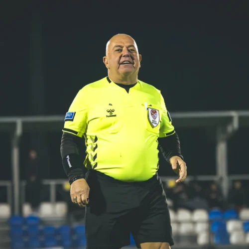 Sędzia piłkarski w jaskrawej koszulce oraz czarnych spodenkach na boisku, uśmiechnięty i w dynamicznej pozie.