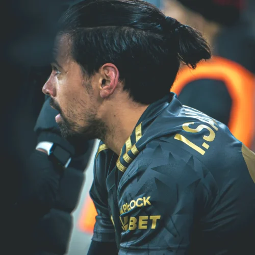 Na zdjęciu widoczny jest piłkarz w szarej koszulce z złotymi detalami, skupiony i siedzący na ławce rezerwowych w czasie meczu.