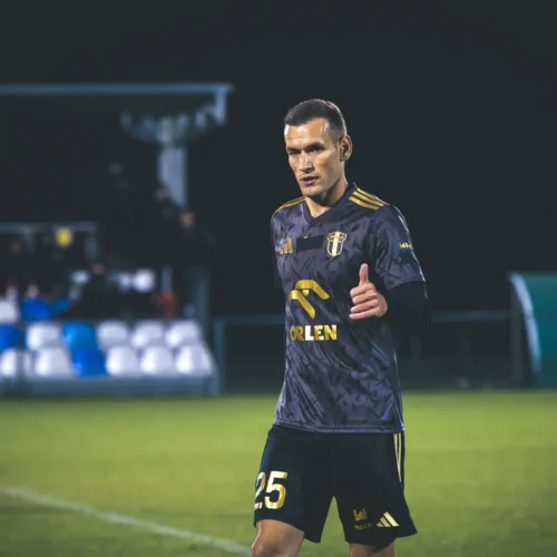 Na zdjęciu widoczny jest piłkarz w czarnym stroju sportowym, który macha kciukiem w górę na boisku podczas meczu.