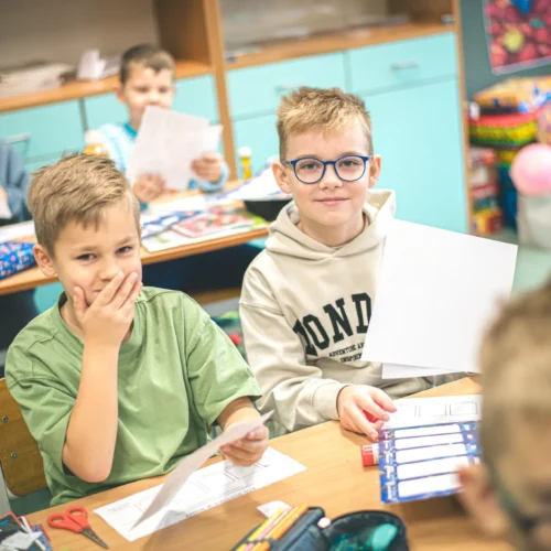Na zdjęciu w klasie siedzą uczniowie, z których dwóch uśmiecha się, trzymając kartki papieru, podczas gdy w tle widać innych chłopców zajętych pracą.