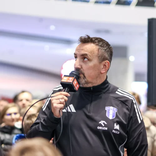 Mężczyzna w czarnej bluzie sportowej z logo klubu mówi do mikrofonu wśród zgromadzonego tłumu.