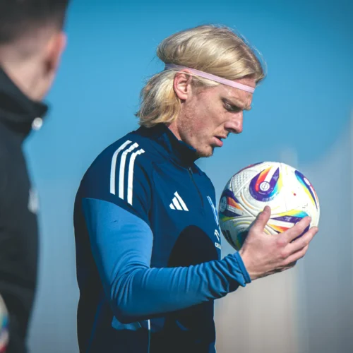 Na zdjęciu znajduje się zawodnik piłkarski trzymający piłkę, skoncentrowany i zamyślony, w trakcie treningu na boisku.