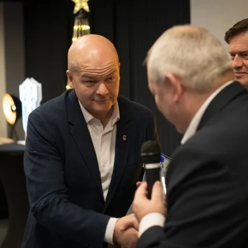 Na zdjęciu widoczna jest scena z uroczystości, gdzie dwóch mężczyzn ściska sobie dłonie, a w tle znajdują się dekoracje świąteczne oraz stół z poczęstunkiem.