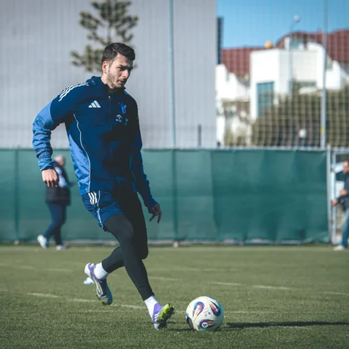 Piłkarz w niebieskim stroju trenuje na boisku, kopiąc piłkę na zielonej murawie w słoneczny dzień.