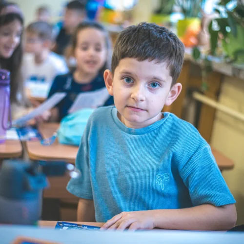 Na zdjęciu widać chłopca w niebieskiej koszulce siedzącego przy biurku w klasie, przed którym znajdują się uczniowie w tle.