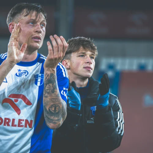 Na zdjęciu widać dwóch mężczyzn w niebiesko-białych strojach, którzy klaskają i wyrażają emocje po meczu, znajdując się na stadionie.