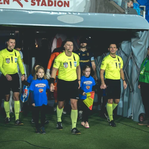 Na zdjęciu sędziowie piłkarscy w żółtych strojach wychodzą na boisko, trzymając za ręce dzieci w niebieskich koszulkach.