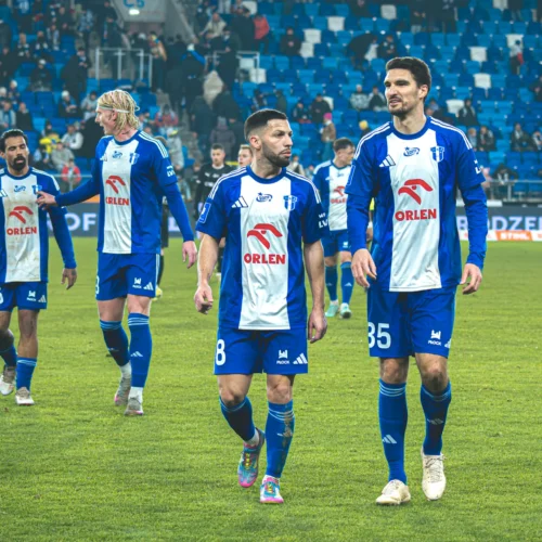 Na zdjęciu widać czterech piłkarzy, którzy spacerują po boisku, ubrani w niebieskie stroje z białymi elementami i logo sponsora.