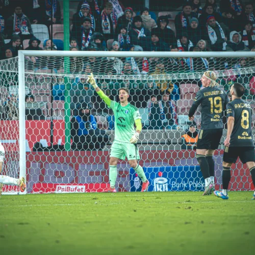 Na zdjęciu widać mecz piłkarski z bramkarzem, który interweniuje w obronie bramki, a w tle znajdują się kibice oraz zawodnicy obu drużyn.