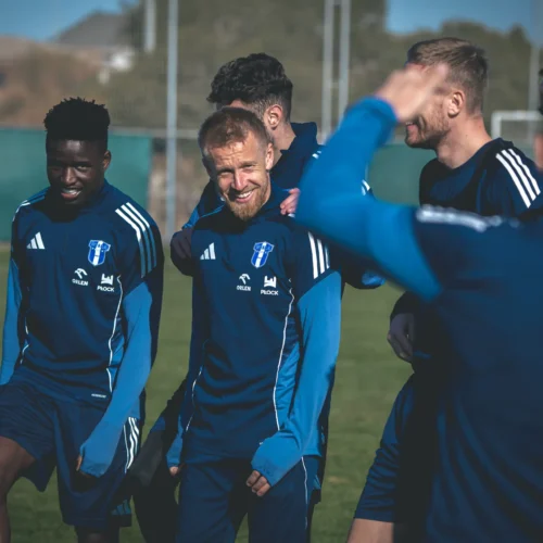 Grupa piłkarzy w niebieskich strojach treningowych uśmiecha się i rozmawia na boisku podczas treningu.