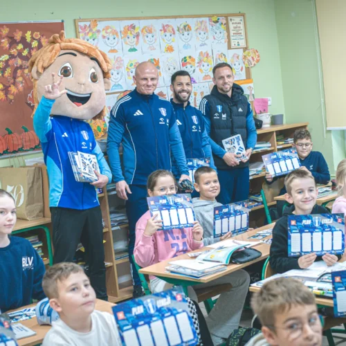 Zdjęcie przedstawia dzieci w klasie, które trzymają kolorowe plany zajęć, w towarzystwie trzech mężczyzn oraz maskotki w kształcie chłopca.