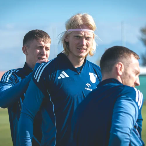 Na zdjęciu trzej mężczyźni w niebieskich strojach sportowych rozmawiają ze sobą na boisku treningowym.