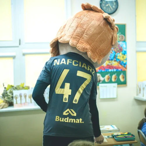 Na zdjęciu widać postać w kostiumie mascota z dużą głową, ubraną w koszulkę sportową z numerem 47, stojącą w klasie obok uczniów.