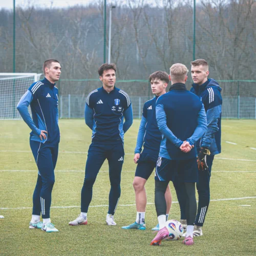 Pięciu zawodników futbolowych stoi na boisku, rozmawiając ze sobą w sportowych strojach treningowych.