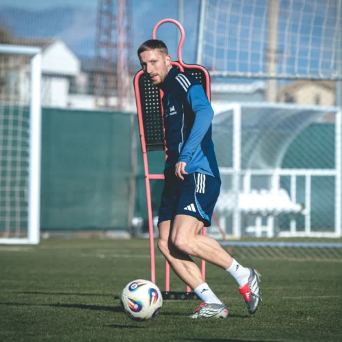 Mężczyzna w niebieskiej koszulce trenuje piłkę nożną na boisku, z piłką obok niego i bramką w tle.