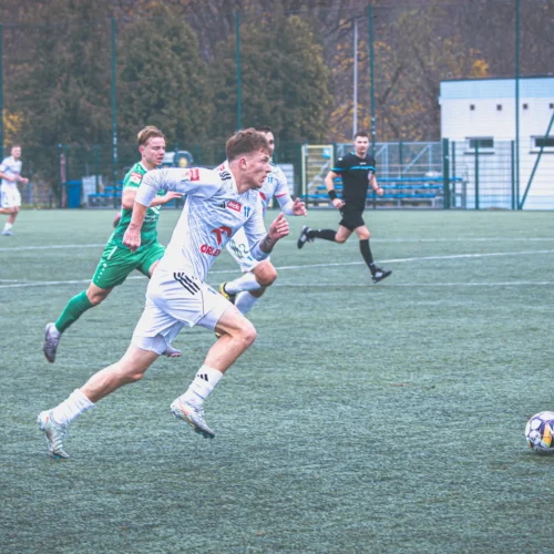 Na zdjęciu widać zawodników piłki nożnej biegnących w stronę piłki na boisku trawiastym.