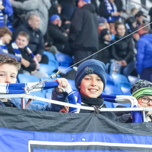 Trzech uśmiechniętych chłopców trzymających transparent na stadionie, wśród tłumu kibiców.