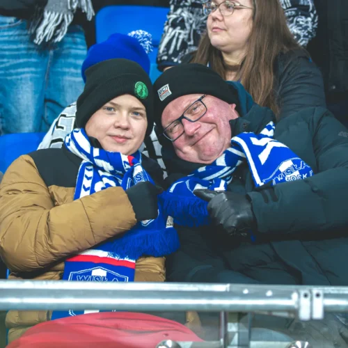 Na zdjęciu dwóch kibiców siedzi na trybunach, ubranych w szaliki w kolorach ich drużyny, uśmiechając się i pozując do zdjęcia.