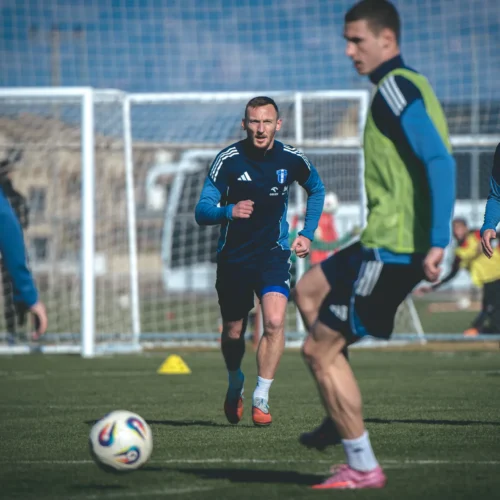 Zdjęcie przedstawia grupę piłkarzy trenujących na boisku w słoneczny dzień, z jednym zawodnikiem wyraźnie biegnącym w kierunku piłki.