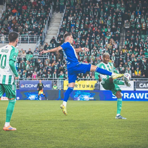 Zawodnicy rywalizują na boisku podczas meczu piłkarskiego, gdzie jeden z graczy unosi nogę, aby wykonać szarżę na przeciwnika.