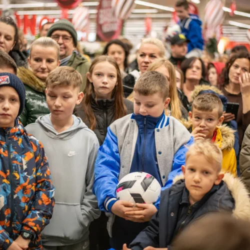 Grupa dzieci i dorosłych w zatłoczonej przestrzeni, wśród których jedno z dzieci trzyma piłkę nożną, a inne mają skupione wyrazy twarzy.