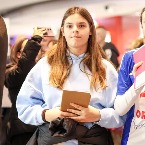 Na zdjęciu widać młodą dziewczynę w jasnoniebieskiej bluzie trzymającą notatnik, stojącą w tłumie ludzi.