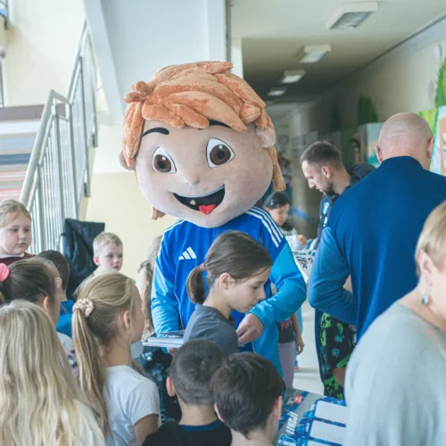 Na zdjęciu widoczny jest clown w wielkiej masce z uśmiechniętą twarzą, który wita grupę dzieci w szkolnym korytarzu.