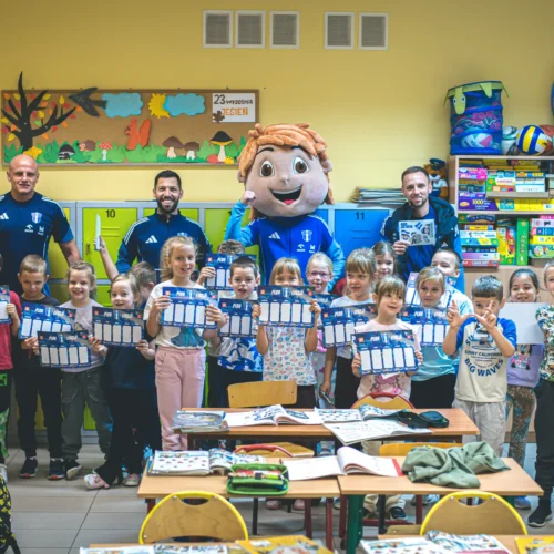 Na zdjęciu widoczna jest grupa dzieci w klasie, które wraz z dwoma trenerami sportowymi oraz maskotką trzymają certyfikaty, a w tle widać kolorowe meble szkolne.