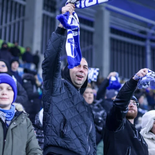 Na zdjęciu widać grupę kibiców sportowych, którzy z entuzjazmem trzymają w rękach szaliki, wyrażając radość podczas wydarzenia na stadionie.