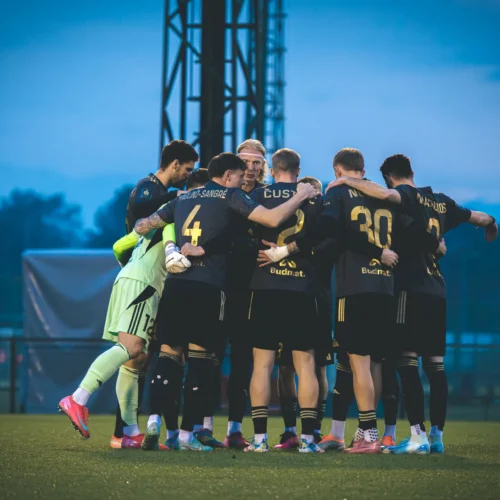Zespół piłkarski zebrany w kręgu przed meczem, w tle niebo w odcieniach błękitu.