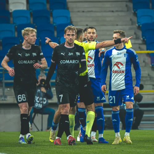 Na zdjęciu widać grupę zawodników piłkarskich na boisku, podczas meczu, z wyraźnymi oznaczeniami drużyn.