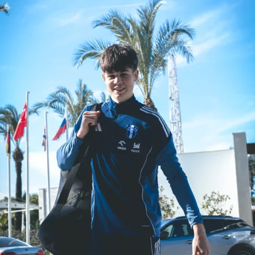 Na zdjęciu młody sportowiec w niebieskim stroju stoi z uśmiechem, trzymając torbę sportową i czerwoną torbę na tle palm oraz flag krajów.