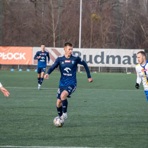 Na zdjęciu występuje mecz piłkarski, w którym zawodnik w niebieskim stroju biegnie z piłką, a dwóch przeciwników w białych stroje o odcieniach niebieskiego goni go na boisku.