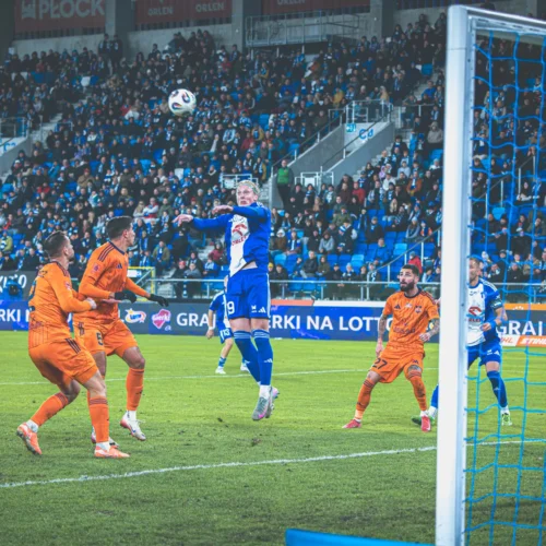 Na zdjęciu widać zawodnika skaczącego do piłki podczas meczu piłki nożnej, z rywalizującymi zawodnikami w pomarańczowych strojach oraz tłumem kibiców na trybunach.