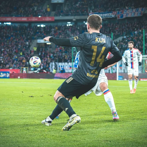 Na zdjęciu widoczna jest intensywna akcja piłkarska, w której zawodnik w czarnym stroju z numerem 14 wykonuje zagranie piłką, a w tle widać tłum kibiców oraz innych graczy.